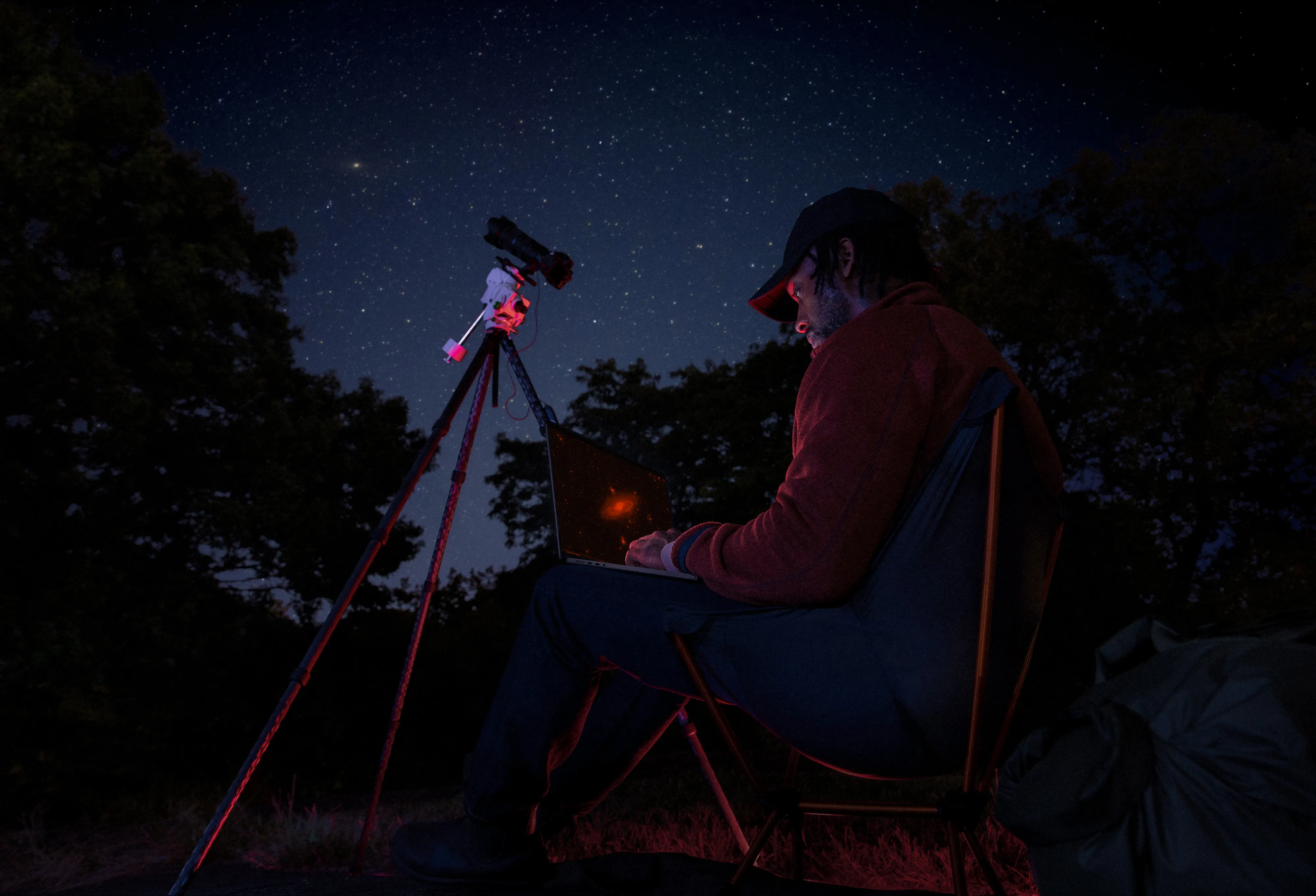 Astronomer using a laptop and telescope outdoors at night to view a galaxy under a starry sky.